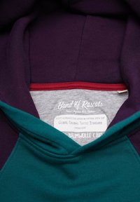 Huva sweatshirt med en färgblockdesign i lila och teal, med en grå etikett med text och röd sömnad vid kragen.