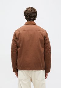 Bărbat cu păr creț de culoare brună, îmbrăcat într-o jachetă maro și pantaloni crem, stând cu fața către un fundal alb simplu.