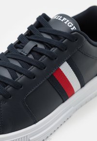 Baskets en cuir marine avec surface texturée, lacets noirs plats, semelle en caoutchouc blanche, et rayures horizontales rouges, blanches et marines sur le côté.