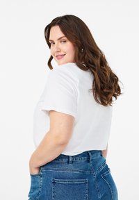 Biele bavlnené tričko s krátkymi rukávmi, nosené s vysokými modrými džínami. Modelka má hnedé vlnité vlasy a je otočená trochu nabok.