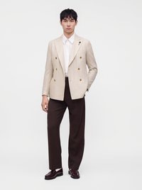 Jeune homme portant un blazer beige croisé, une chemise blanche, un pantalon marron foncé et des mocassins marron foncé, debout sur un fond blanc.