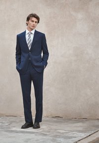 Jeune homme en costume bleu marine, cravate rayée et chaussures noires, debout les mains dans les poches contre un mur beige.