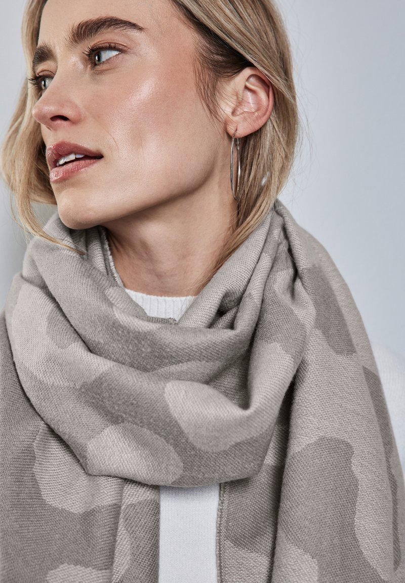 Écharpe grise avec un motif camouflage subtil, fabriquée en matériau doux. Enroulée autour du cou, elle présente une texture lisse et tombe élégamment.