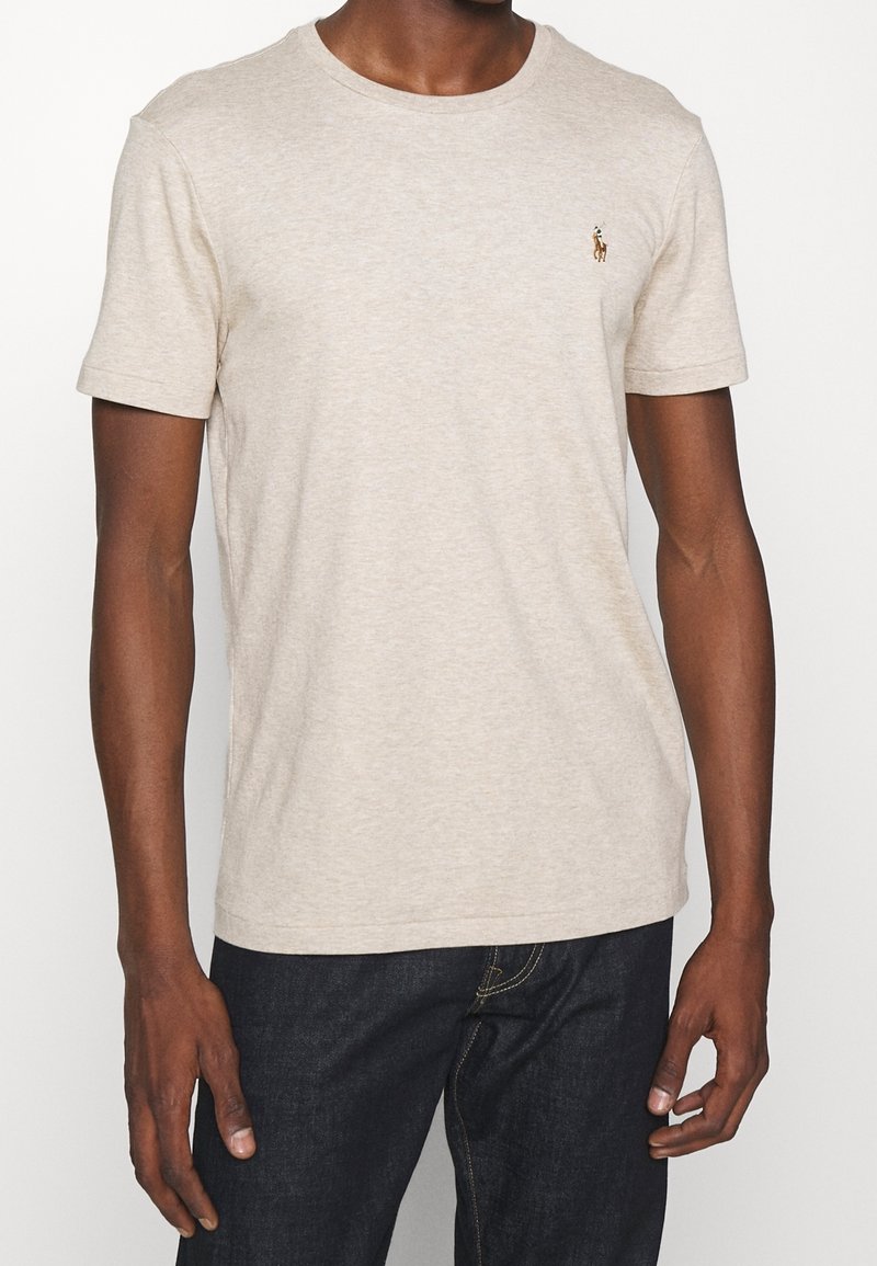Mann in einem beigen kurzärmligen Rundhalsausschnitt-T-Shirt mit kleinem gesticktem Logo auf der Brust und dunkelblauen Jeans.