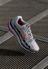 Zapatilla Nike gris con materiales de malla y sintéticos; presenta acentos rosas y amarillos, un logo prominente y una suela texturizada para mayor tracción.