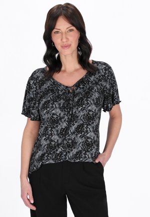 Kvinde med mørkt, bølget hår iført øreringe i ringeform, en sort og grå bluse med blomsterprint og korte ærmer samt sorte bukser, smiler let.