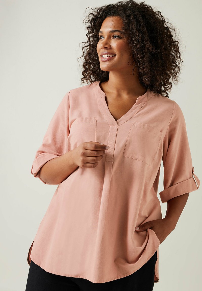 Vaaleanpunainen pusero, jossa on V-pääntie, kaksi rintataskua ja käärityt hihat. Pehmeä kangas, rento malli ja kaareva helma.