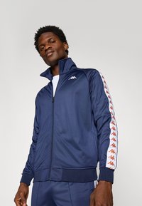Kappa zip-up jakke, marineblå, laget av glatt stoff. Har høy krage, hvit logo og sidestriper med gjentatt rød merkevare.