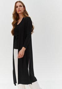 Czarny długi kardigan z długimi rękawami, otwartym przodem i bocznymi rozcięciami. Noszony na czarną bluzkę i białe spodnie. Gładki materiał i luźny krój.