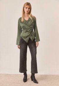 Top verde de manga larga con detalle de nudo en la cintura, combinado con vaqueros de pierna ancha negros con bordado floral y botas de tobillo.