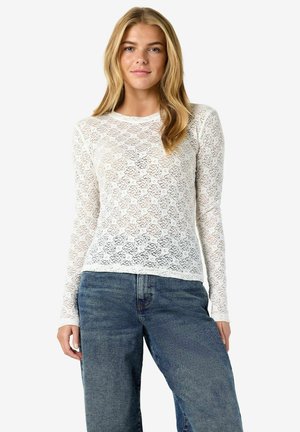 Kvinde iført en hvid langærmet blondebluse og blå jeans, stående og vendt fremad med et neutralt ansigtsudtryk.