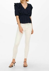Top avvolgente navy con maniche arricciate, tessuto strutturato, abbinato a jeans skinny bianchi e sandali neri con cinturini. Design semplice e pulito.