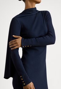 Tamsiai mėlyna ilgomis rankovėmis suknelė su drapiruotu kaklu, pagaminta iš tekstūruotos medžiagos, su aukso spalvos sagomis rankogaliuose. Elegantiška silueto linija su apsiausto detale.