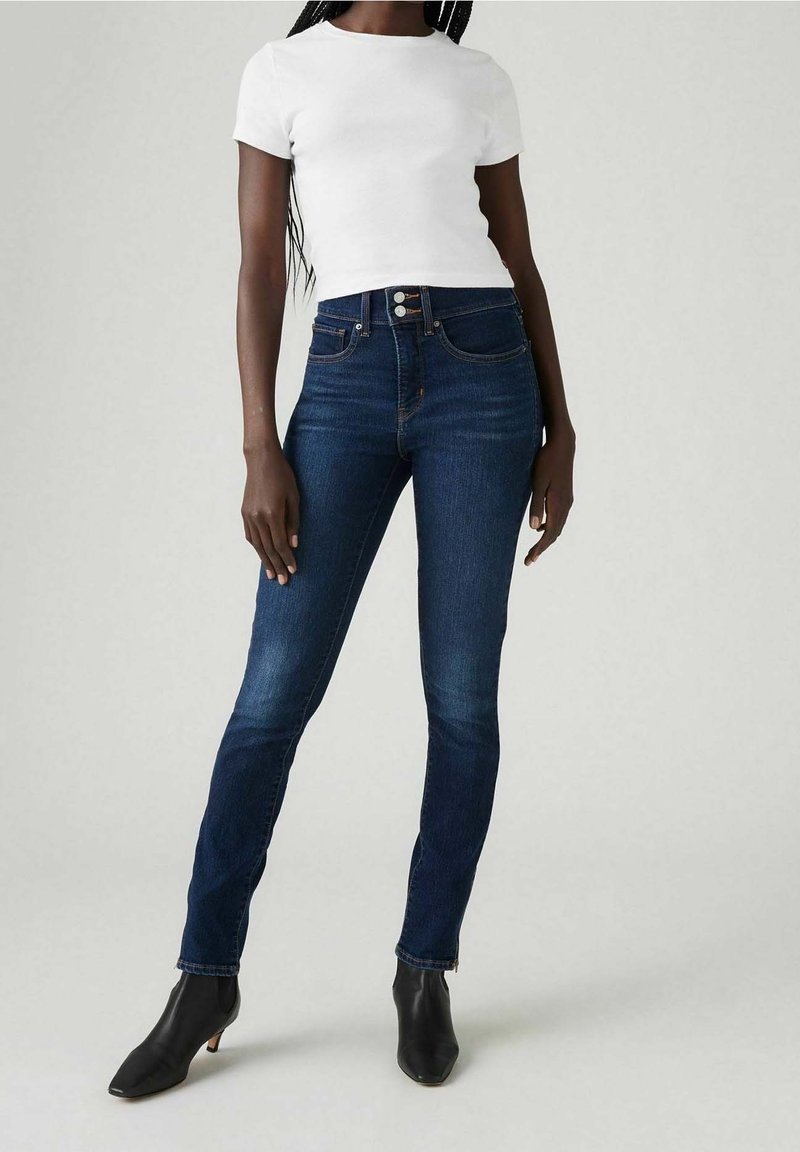 Hubené džíny v tmavě modrém denimu s vysokým pasem, stylizované s bílým upnutým tričkem a černými koženými kotníkovými botami na podpatku. Hladká textura, minimální detaily.