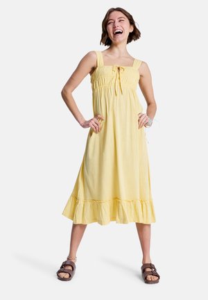 Jeune femme riant, debout avec assurance, les mains sur les hanches, portant une robe midi jaune sans manches et des sandales marron sur fond blanc.