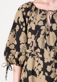 Blouse floral noir et tan en tissu léger ; présente une ouverture en forme de trou de serrure et des manches bouffantes avec des fermetures à nouer au niveau des poignets.