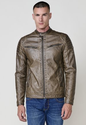 Hombre vistiendo una chaqueta de cuero marrón con bolsillos con cremallera y jeans azules, de pie contra un fondo gris claro.