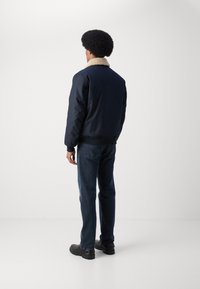 Chaqueta bomber de color marino con un collar de piel sintética crema, puños y dobladillo acanalados, combinada con jeans azules y zapatos negros, vista desde atrás.