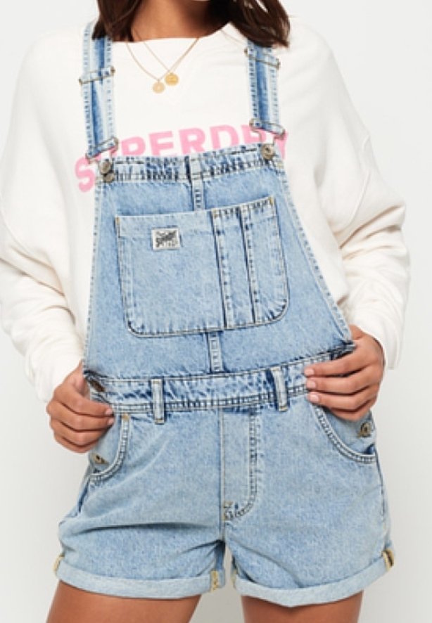 Femme portant une salopette courte en denim bleu clair et un sweat-shirt blanc avec des lettres roses, tenant les côtés de la salopette.