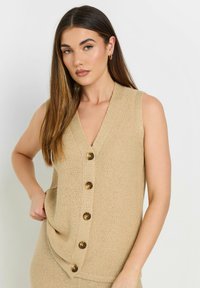 Beige stickad ärmlös väst med V-ringning och fem stora runda knappar fram. Texturerad tyg med sidfickor.