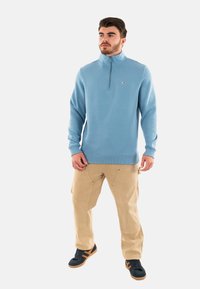 Pullover azzurro chiaro con colletto a mezza zip, maniche e orlo a coste, abbinato a pantaloni cargo beige e scarpe nere con dettagli arancioni.