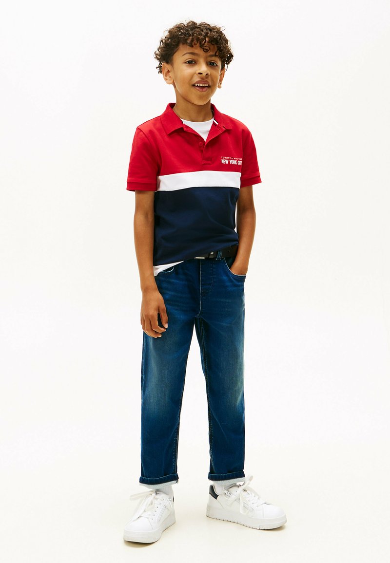 Dreng med krøllet hår iført en rød, hvid og marineblå polo shirt, blå jeans og hvide sneakers, står med den ene hånd i lommen.