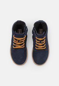 Granatowe zamszowe botki za kostkę z żółtymi sznurowadłami i zamkami błyskawicznymi po bokach. Podeszwy są w odcieniu jasnobrązowym z haftowanym wzorem, a wnętrze jest miękkie.