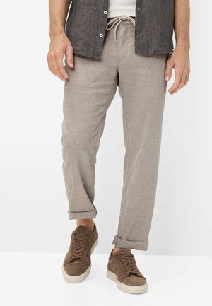 Mann trägt hellbeige Leinenhose mit hochgekrempelten Säumen, ein dunkelgraues Hemd mit Knöpfen und braune Wildleder-Sneaker mit weißen Sohlen.