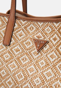 Sac fourre-tout tissé marron et blanc avec des poignées en cuir marron et une plaque triangulaire marron avec le logo Guess USA.