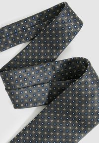 Sötét szürke, geometrikus mintázatú nyakkendő kék és ezüst díszítéssel. Simább textúra, keskeny forma és fokozatos vége.
