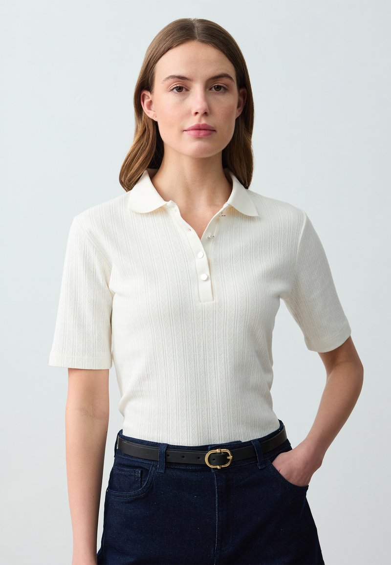Witte polo met korte mouwen en een getextureerd ribbelpatroon, knoopsluiting en een klassieke kraag. Draag het met een donkerblauwe spijkerbroek en een zwarte riem.