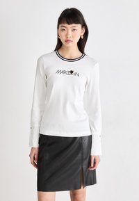 Camisa blanca de manga larga con cuello negro y logo bordado, combinada con una falda mini de cuero negra con una abertura lateral.