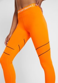 Pomarańczowe legginsy z wysokim stanem z kontrastującymi ciemnymi szwami. Posiadają detale paneli na udach oraz markowy pasek w talii. Gładka tekstura.