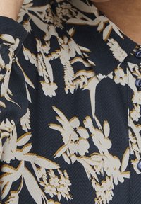 Blommig blus i marinblått med krämfärgade och guldiga mönster, med en strukturerad design, spetsig krage och knäppning.