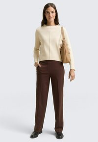 Beżowy sweter z okrągłym dekoltem, długimi rękawami i pionowym szwem. Ciemnobrązowe spodnie taliowane o prostym kroju. Jasnobrązowa torebka na ramię. Skórzane buty.