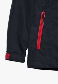 Giacca blu navy con zip rossa, tessuto testurizzato e polsino regolabile con strappo rosso in velcro. Dettaglio ravvicinato della manica.