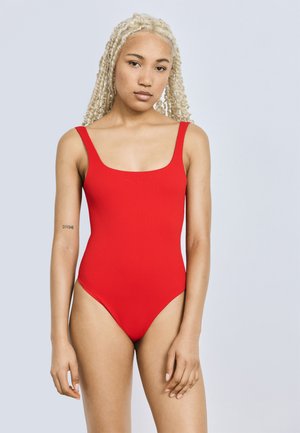 Kvinne med lange blonde fletter iført en rød badedrakt, stående mot en lys grå bakgrunn med en "DIVINE" tatovering på venstre arm.