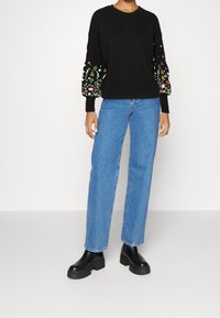 Pull noir en tricot avec broderie florale colorée sur les manches, associé à un jean bleu à jambes droites et des bottes noires à semelles épaisses.