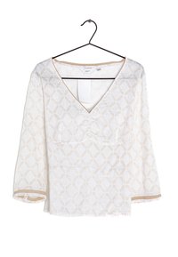 Blusa blanca con patrón geométrico beige, escote en V y mangas de tres cuartos colgando de una percha negra sobre un fondo blanco.