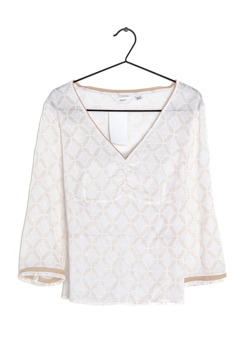 Blusa blanca con patrón geométrico beige, escote en V y mangas de tres cuartos colgando de una percha negra sobre un fondo blanco.