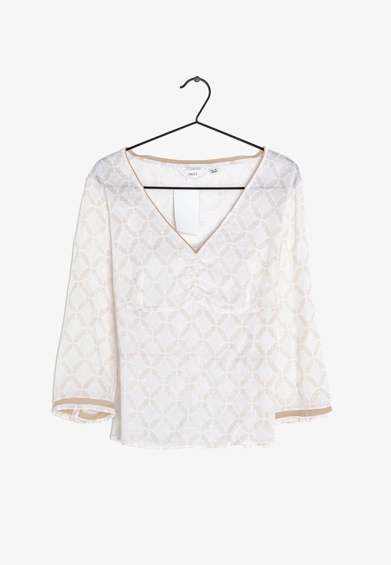 Blusa blanca con patrón geométrico beige, escote en V y mangas de tres cuartos colgando de una percha negra sobre un fondo blanco.