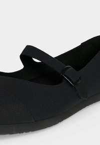 Chaussure plate Mary Jane noire texturée avec bride réglable et bout arrondi sur fond blanc.
