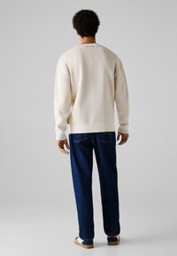 Cremefarbener Pullover mit gerippten Bündchen und Saum, kombiniert mit dunkelblauen Jeans. Die Rückansicht zeigt das Logodetail am Halsauschnitt und legere Schuhe.
