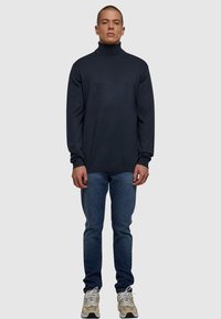 Mørkeblå høyhalset genser i myk stoff, avslappet passform, lange ermer, kombinert med mørkeblå jeans og beige joggesko.