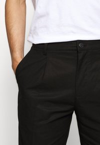 Mužský torzo oblečený v černých kalhotách, s jednou rukou v kapse a bílým tričkem přitaženým dovnitř, zobrazené na jednoduchém pozadí.