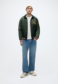 Πράσινο σατέν μπουφάν bomber με κεντητό λογότυπο, λευκό μπλουζάκι, ανοιχτό μπλε τζιν παντελόνι με φαρδιά πόδια και κρεμ sneakers με μαύρες λεπτομέρειες.