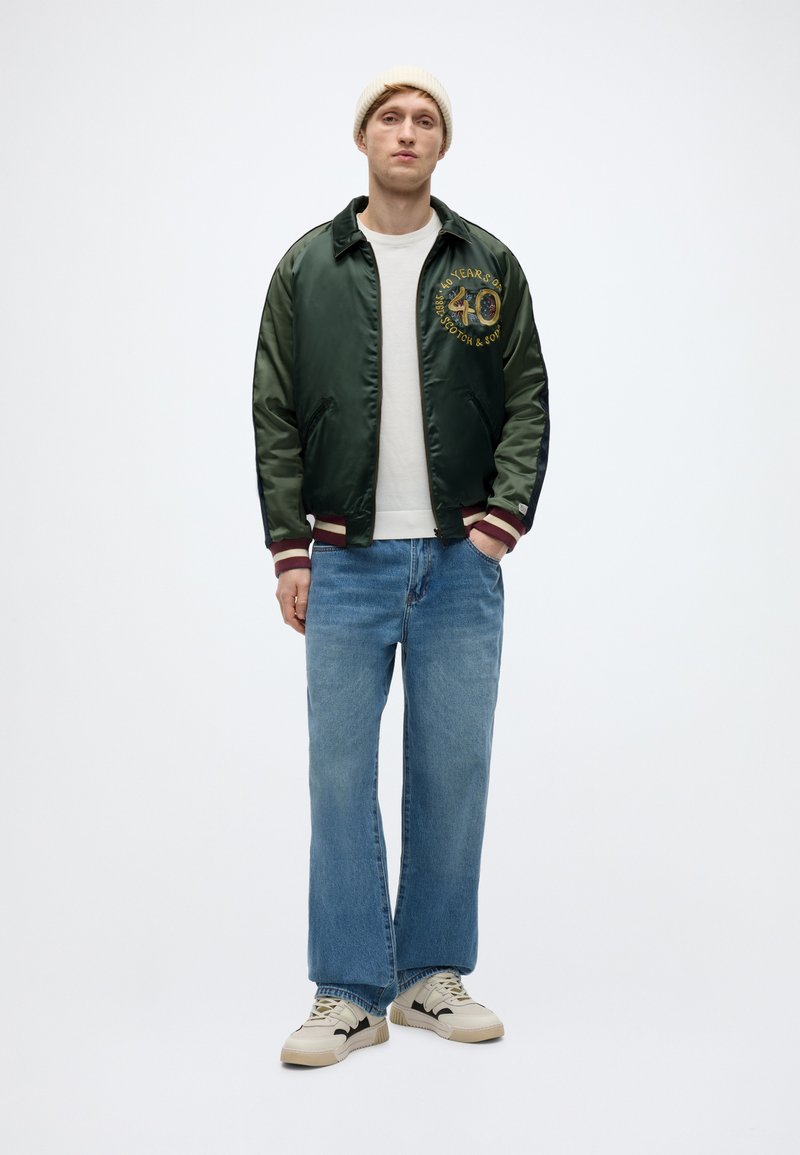 Πράσινο σατέν μπουφάν bomber με κεντητό λογότυπο, λευκό μπλουζάκι, ανοιχτό μπλε τζιν παντελόνι με φαρδιά πόδια και κρεμ sneakers με μαύρες λεπτομέρειες.