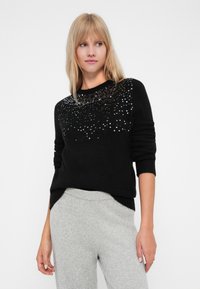 Maglione nero con parte superiore testurizzata caratterizzata da paillettes argentate sparse, maniche lunghe e una vestibilità rilassata, abbinato a pantaloni grigi chiari.