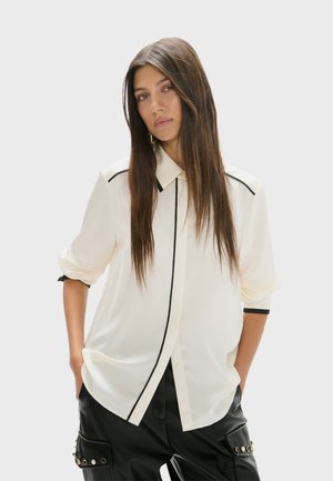 Femme aux longs cheveux bruns vêtue d'un chemisier blanc ample avec une bordure noire et d'un pantalon noir avec des boutons décoratifs, debout les mains dans les poches.