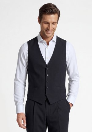 Man draagt een zwart geknoopt vest over een wit overhemd, met bijpassende zwarte broek, glimlacht en kijkt naar beneden.